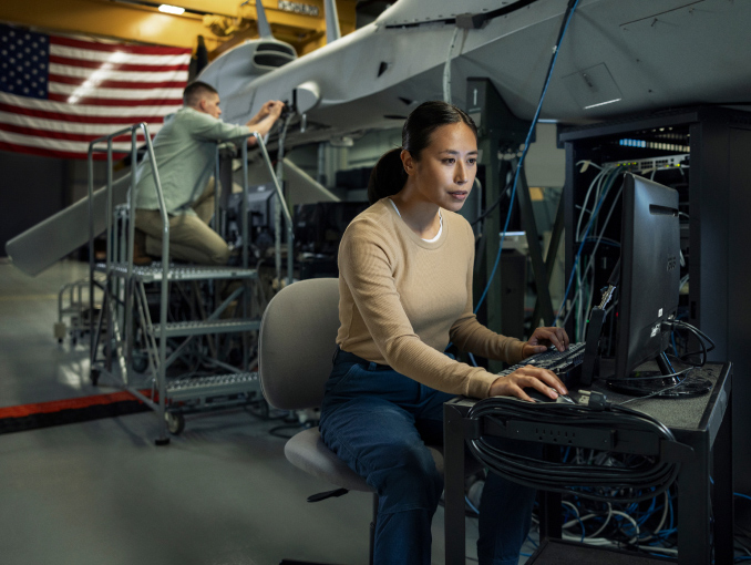 A woman sits at a computer station as a man works on an unmanned aerial vehicle